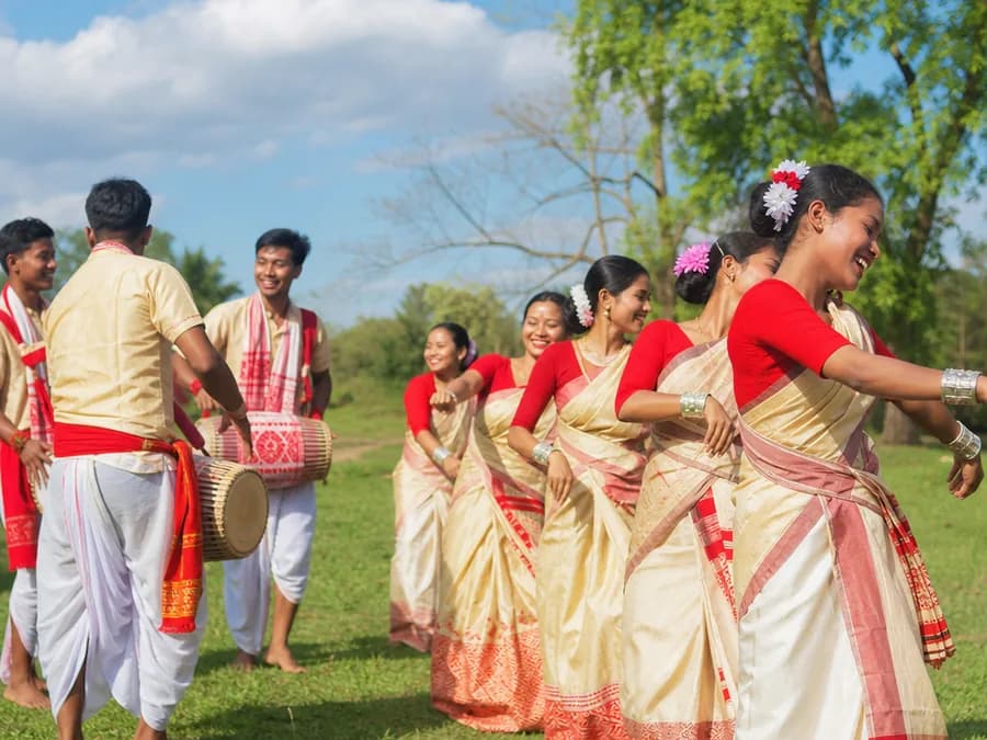 Dancers in traditional red-and-white Assamese dress mid-movement at an outdoor Bihu gathering in bright daylight