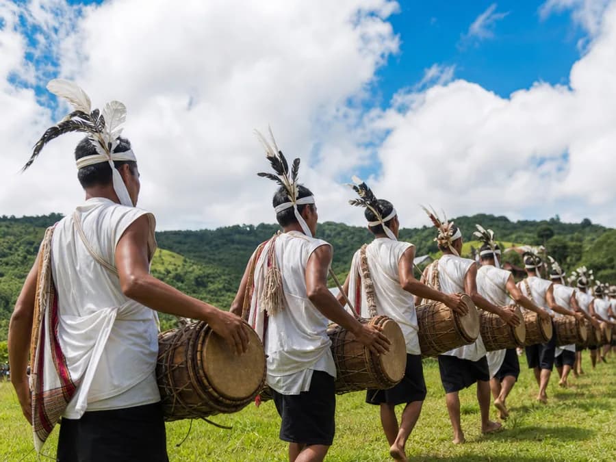 A line of drummers in traditional dress playing long drums at an outdoor Garo harvest festival
