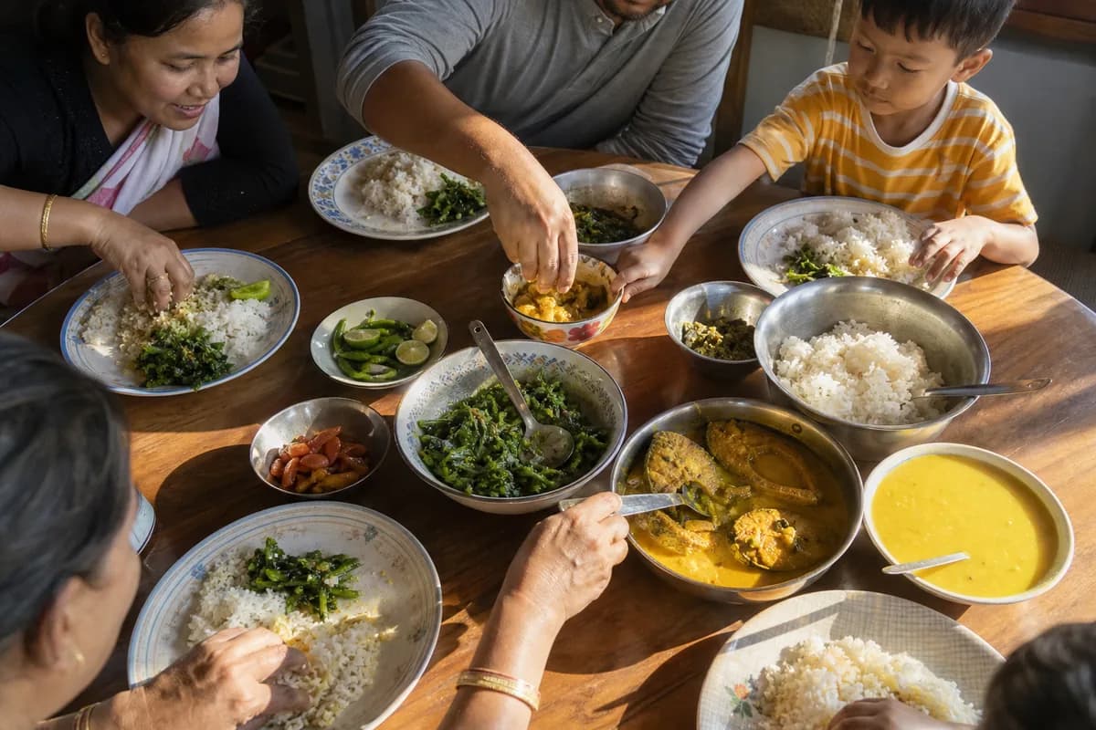 Hands reaching across a sunlit wooden table spread with a home-style Assamese meal of rice, curries and greens