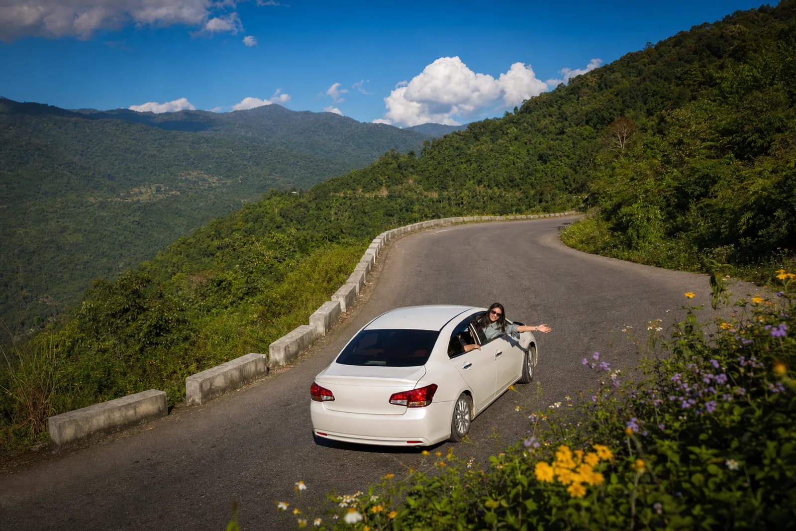 A clean white car on a sunlit hill highway curving between bright green forested slopes