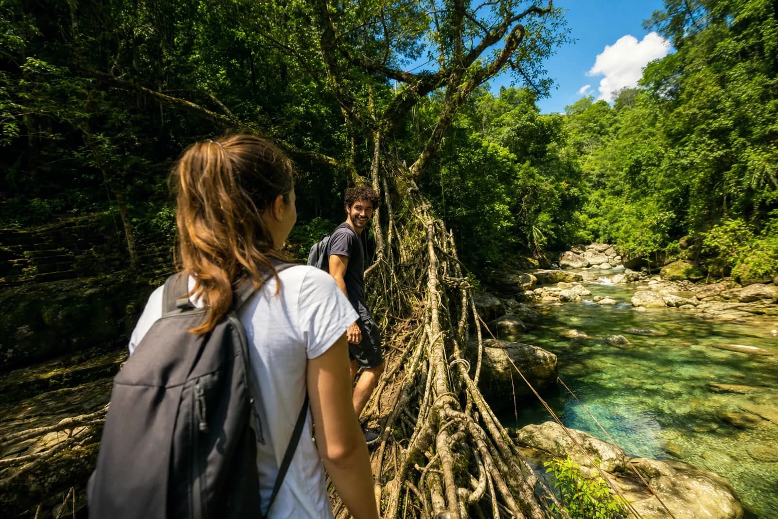 Two young travellers crossing a living root bridge over a clear turquoise stream in sunlit forest